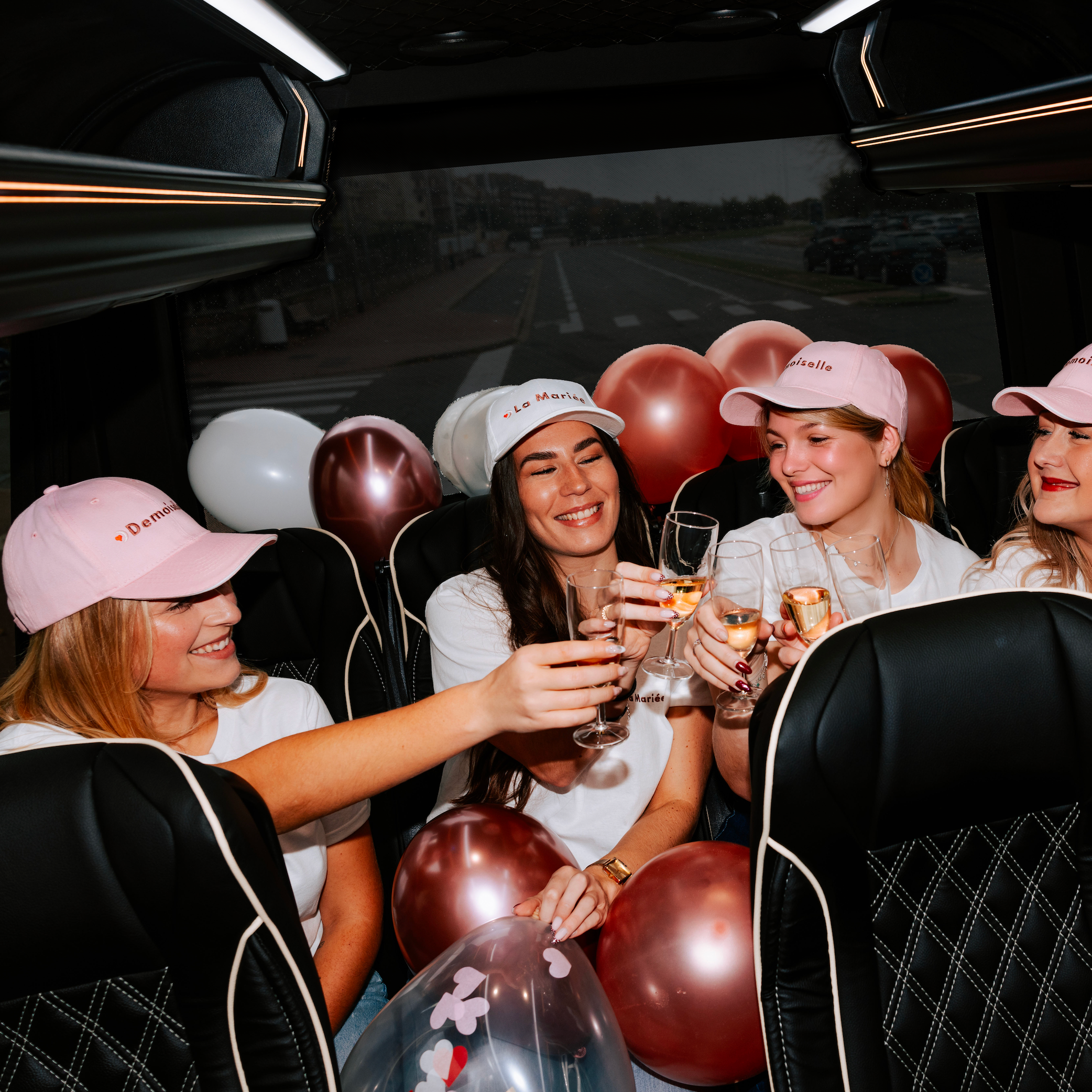 Groupe de femmes portant des casquettes et t-shirts EVJF ‘La Mariée’ et ‘Demoiselle’, trinquant dans un van décoré de ballons lors d’un enterrement de vie de jeune fille.