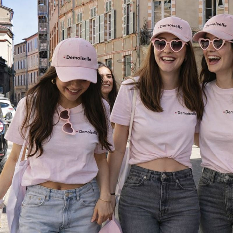Groupe de femmes lors d’un EVJF en ville, portant des t-shirts roses et des casquettes assorties “Demoiselle”, avec des lunettes de soleil en forme de cœur.