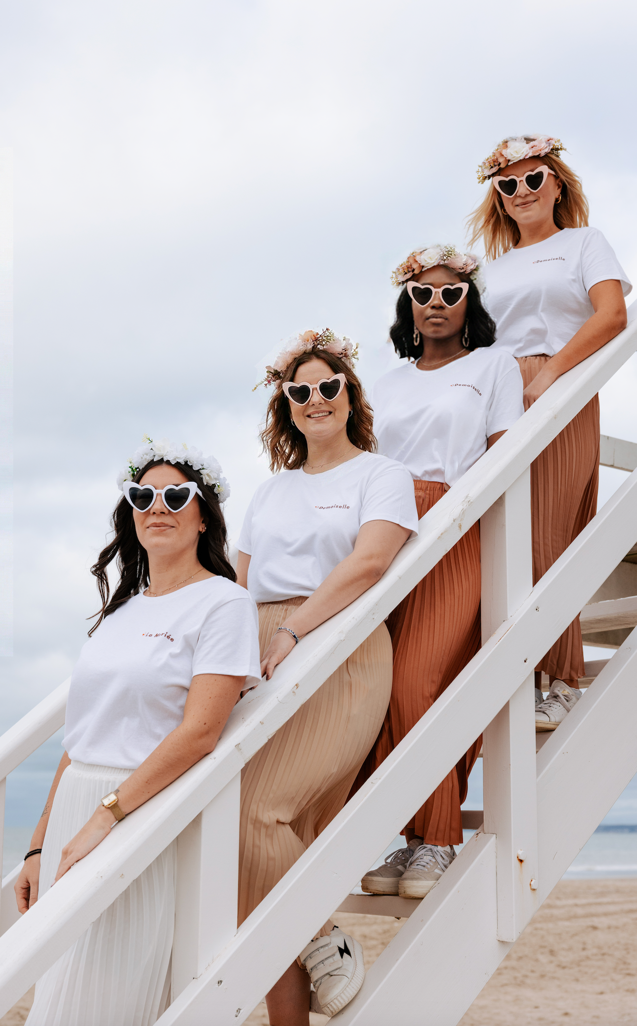 Groupe de femmes lors d’un EVJF, portant des t-shirts blancs assortis, des jupes plissées et des lunettes de soleil en forme de cœur, posées sur un escalier face à la plage.