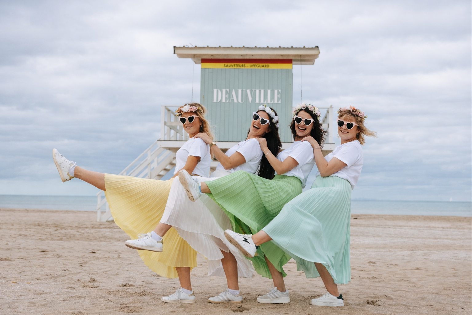 Groupe de femmes portant des t-shirts EVJF, jupes plissées colorées, lunettes cœur et couronnes de fleurs, posant sur la plage devant le poste de secours de Deauville lors d’un enterrement de vie de jeune fille.