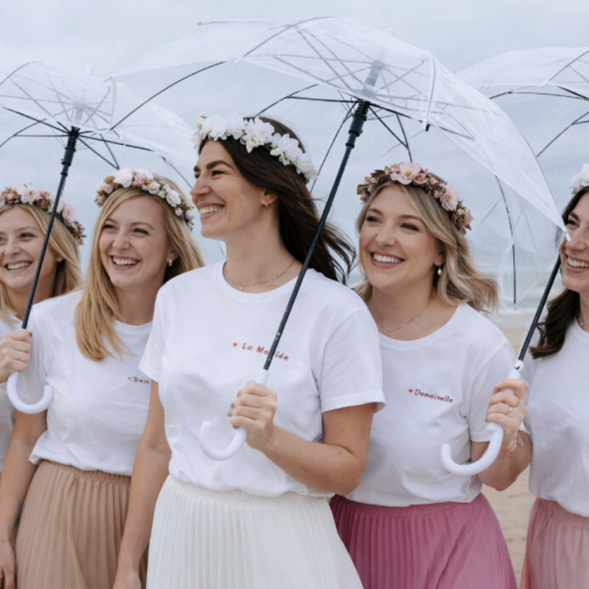 Groupe damies celebrant un EVJF avec des parapluies transparents, tenues assorties et couronnes de fleurs, ambiance joyeuse en exterieur.