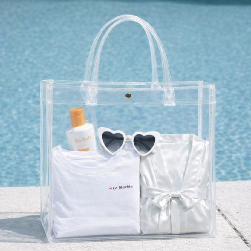 Sac transparent EVJF pose au bord dune piscine avec t shirt future mariee, lunettes coeur et accessoires, idee cadeau pour enterrement de vie de jeune fille.