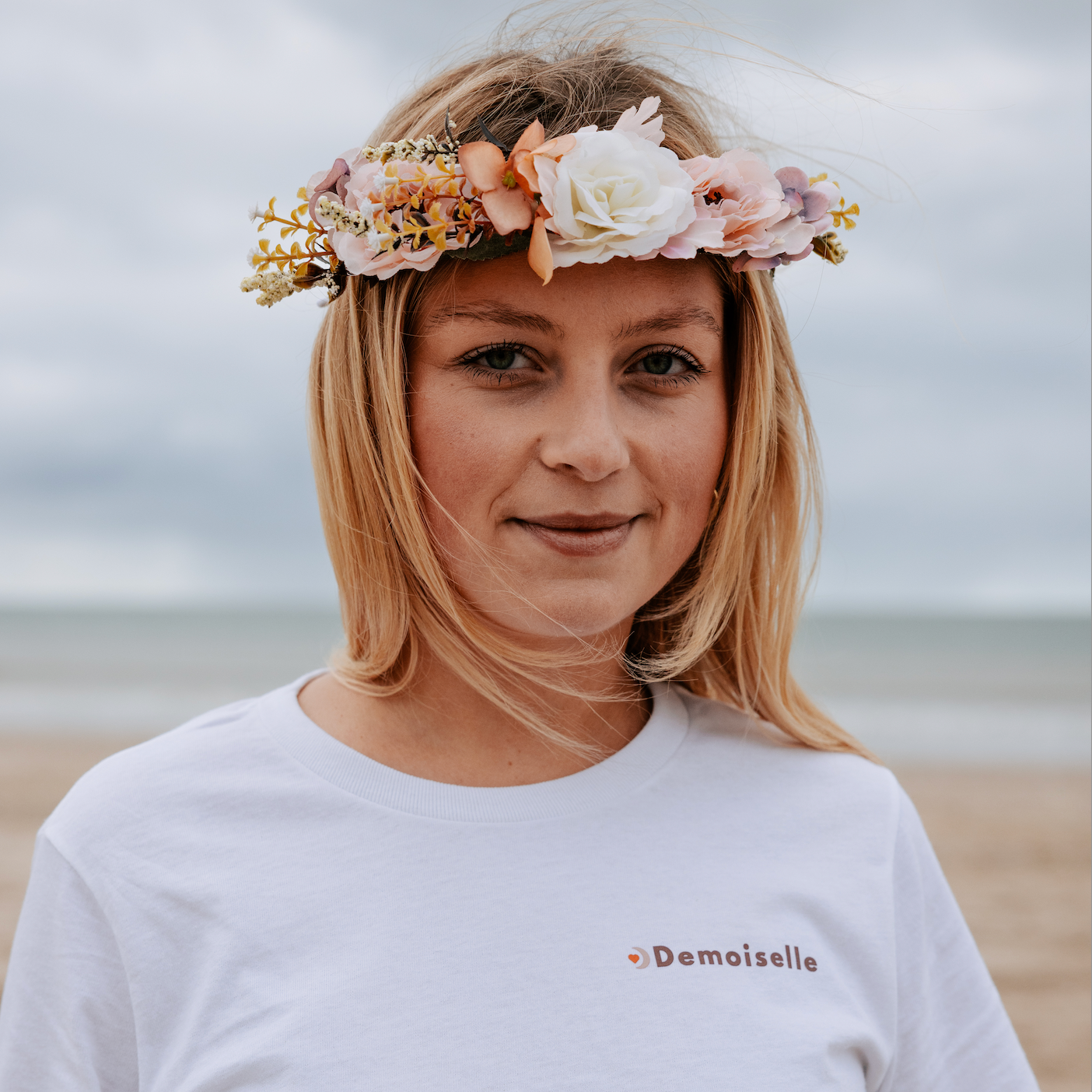 Femme portant un t-shirt blanc EVJF avec inscription ‘Demoiselle’ et une couronne de fleurs, sur la plage, ambiance douce et naturelle pour un enterrement de vie de jeune fille.
