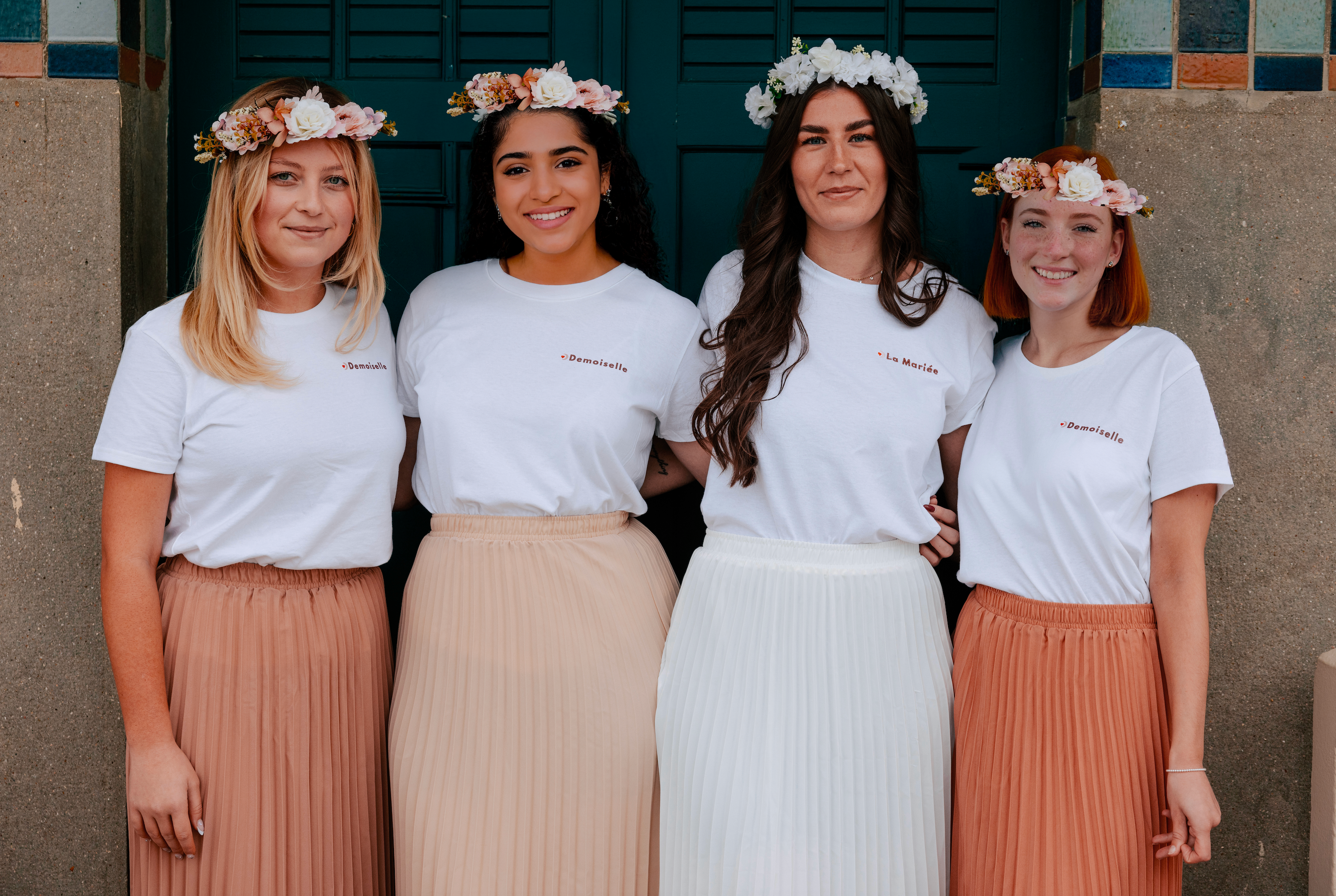 Groupe de femmes portant des t-shirts EVJF ‘La Mariée’ et ‘Demoiselle’ avec des couronnes de fleurs, tenues coordonnées pour un enterrement de vie de jeune fille.