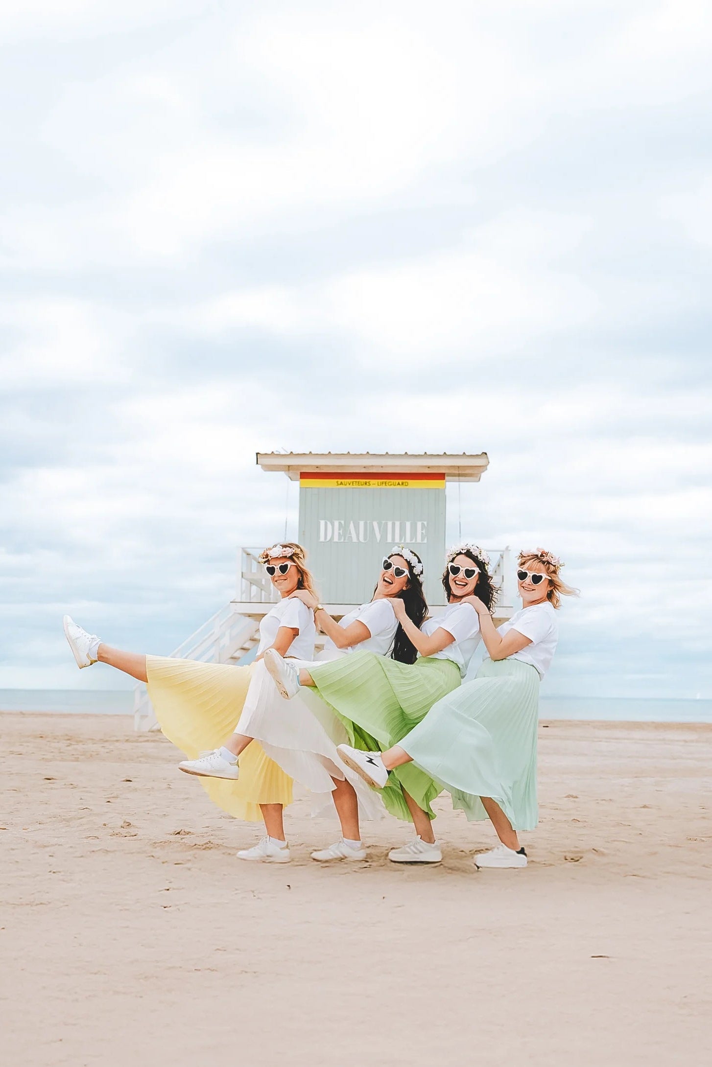 Groupe damies celebrant un EVJF a la plage, portant des jupes plissees colorees et des t shirts blancs, ambiance joyeuse et estivale.