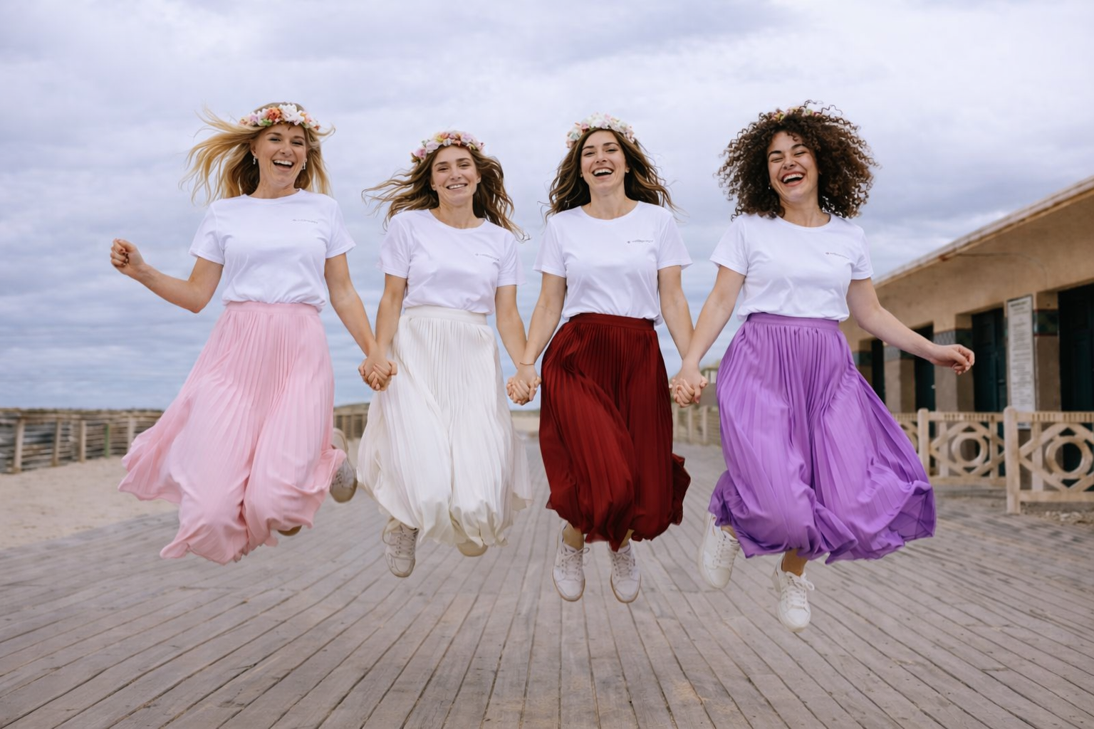 Groupe de femmes portant des t-shirts blancs EVJF et des jupes longues plissées colorées, sautant main dans la main sur une promenade en bord de mer lors d’un enterrement de vie de jeune fille.