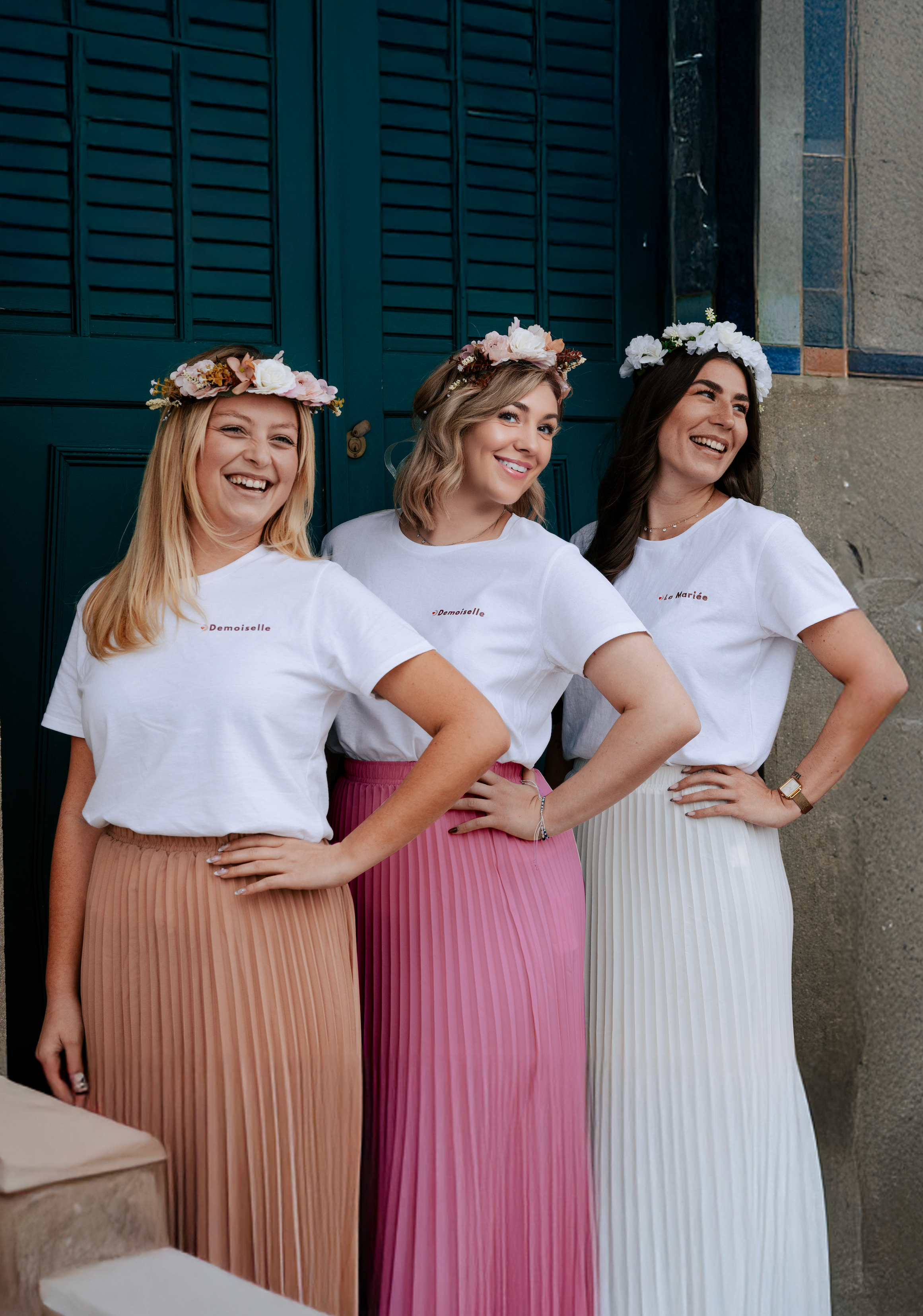 Trois femmes portant des t-shirts EVJF et des jupes longues plissées colorées avec des couronnes de fleurs, posant ensemble lors d’un enterrement de vie de jeune fille.
