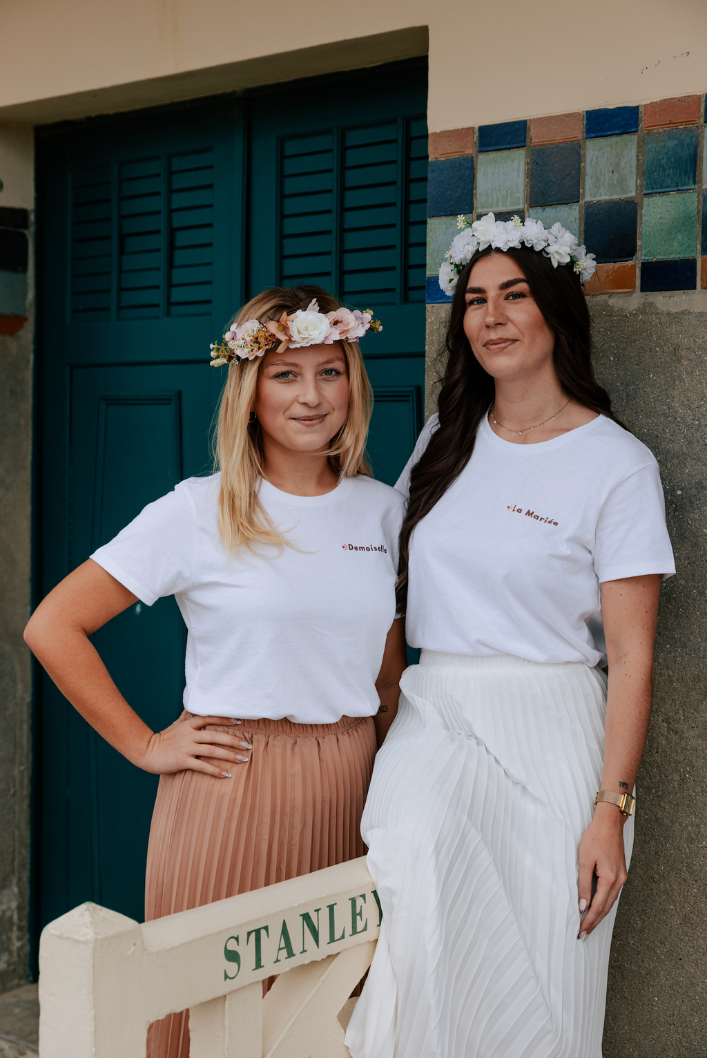 Deux femmes portant des t-shirts EVJF ‘La Mariée’ et ‘Demoiselle’ avec des couronnes de fleurs, tenues coordonnées pour un enterrement de vie de jeune fille.